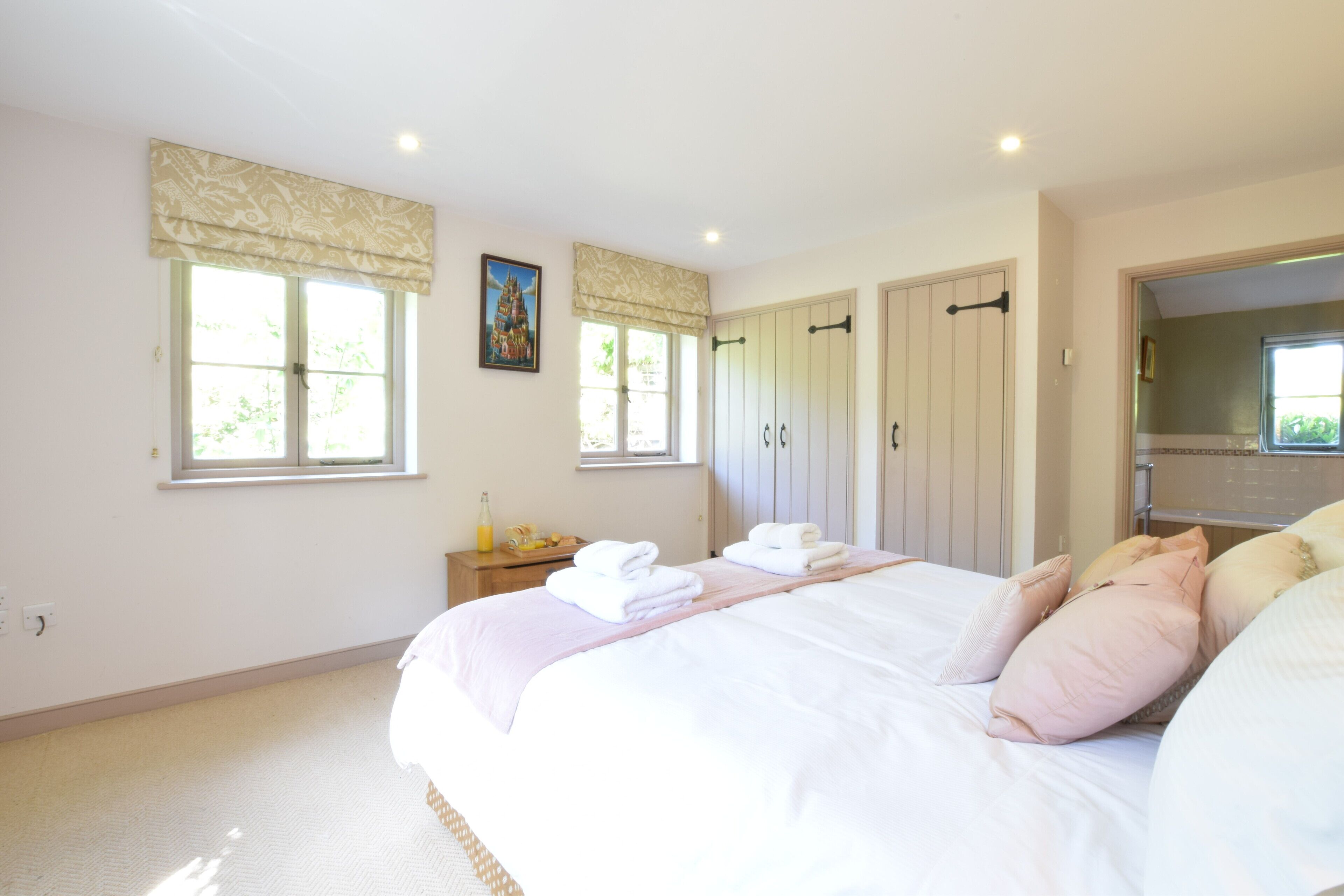 A bedroom with a large bed, two windows with curtains, a bedside table with a bottle, and a mirror.