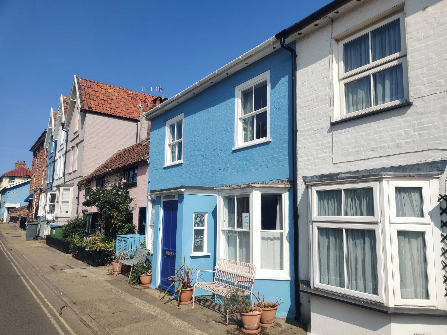 A row of terraced houses with distinct blue and white facades, a clear sky, and a street with a curb and a bench.