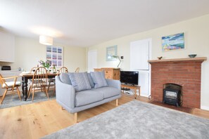 A living room with a sofa, TV, fireplace, dining table, chairs, and a kitchen in the background. - Seaflower, Aldeburgh (Aldeburgh)