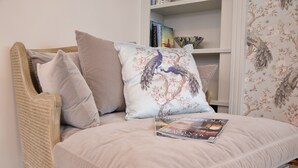 Un llit amb un coixí floral i una revista, una cadira de vimet i una prestatgeria amb llibres i objectes decoratius.