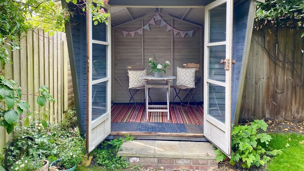 A wooden shed with open doors, a small table with two chairs, and a potted plant. - Rosemary Cottage, Aldeburgh (Aldeburgh)