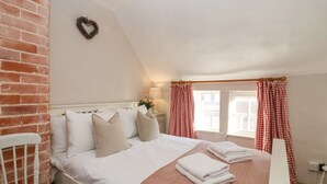 A bedroom with a white bed, a brick wall, a small window with checkered curtains, and a white nightstand. - Rosemary Cottage, Aldeburgh (Aldeburgh)