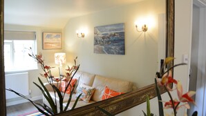 A living room with a beige sofa, a painting on the wall, and a large mirror reflecting the room. - Church Farm Cottage, Dunwich (Saxmundham)