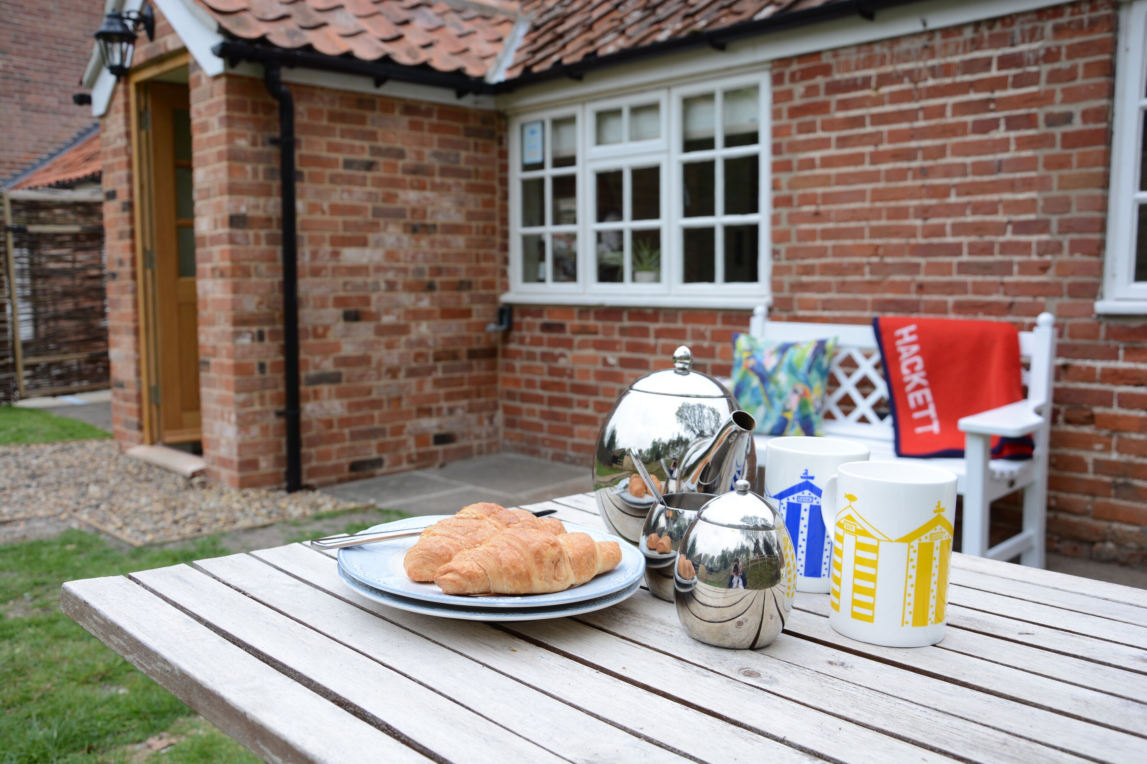 A picnic setup with a croissant on a plate, silver teapot, and two mugs on a wooden table outside a brick house with a window and a door.