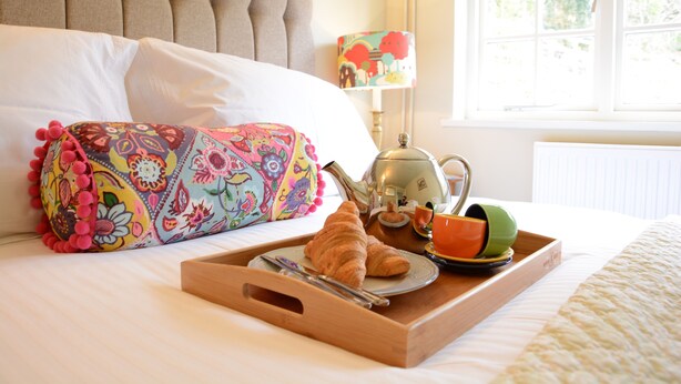 A neatly made bed with a colorful pillow, a tray with croissants, a teapot, and a cup. - Church Farm Cottage, Dunwich (Saxmundham)