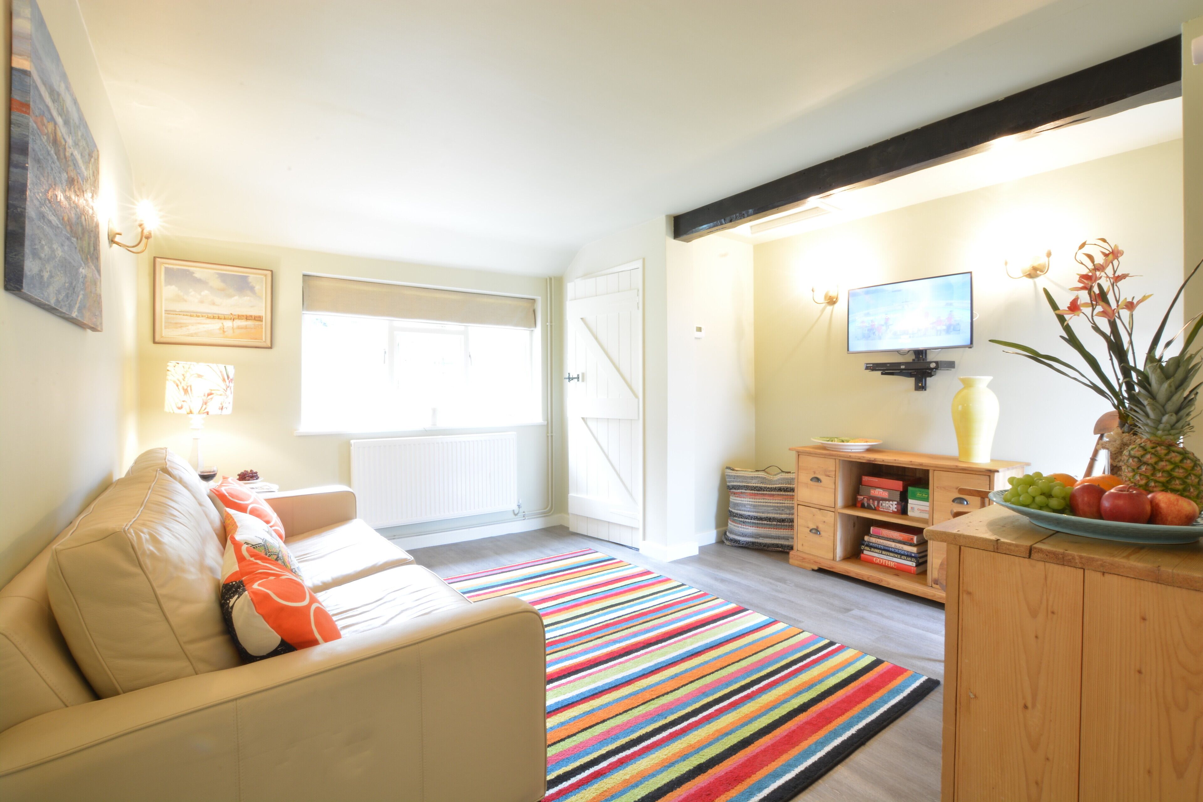 A living room with a beige sofa, a colorful striped rug, a wooden cabinet with fruit, and a television on the wall.