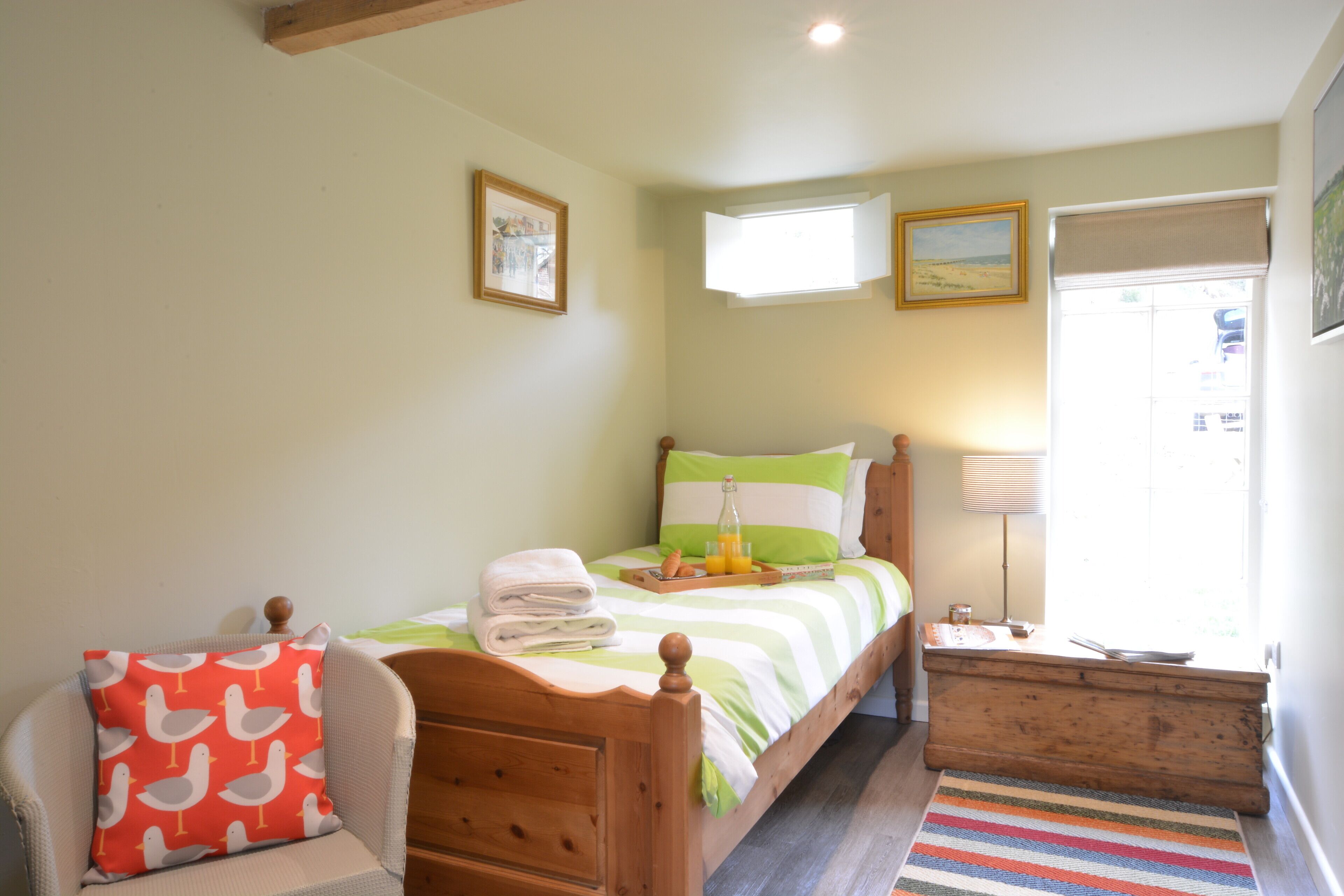 A bedroom with a wooden bed, a chair with a red cushion, a bedside table with a lamp, and a window with a view.