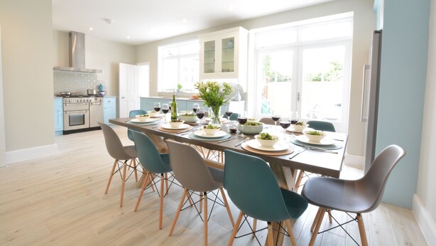 Une cuisine moderne avec un coin repas, comprenant une longue table dressée pour six personnes, des meubles de couleur claire et une grande fenêtre laissant entrer la lumière naturelle.