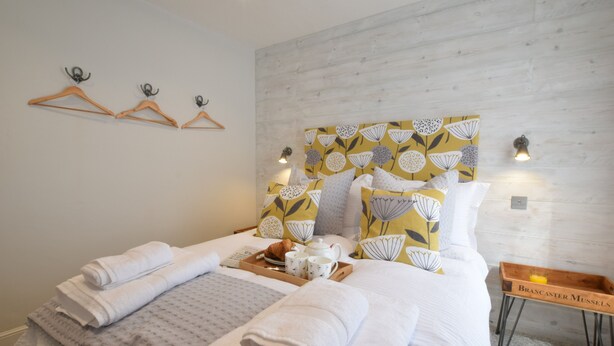 A neatly made bed with towels, a tray with a croissant, and a tray with a drink. There are wooden hangers on the wall and a small table with a sign. - 53 Tern Cottage, Aldeburgh (Aldeburgh)