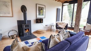 Un salon chaleureux avec un canapé bleu, un poêle à bois noir, un tableau encadré et une vue dégagée sur l’extérieur grâce à une grande fenêtre.