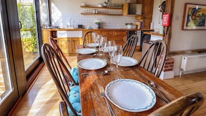 Un coin repas avec une table en bois dressée pour six personnes, entourée de chaises, dans une cuisine dotée de poutres apparentes et offrant une vue à travers une grande fenêtre.