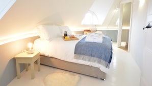 Un dormitorio con techo inclinado, una cama con una bandeja de desayuno, una lámpara de noche y un almohadón blanco mullido.