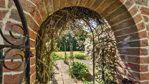 Un passage voûté en briques, surmonté d’une grille métallique, mène à un jardin agrémenté d’un sentier et de verdure.