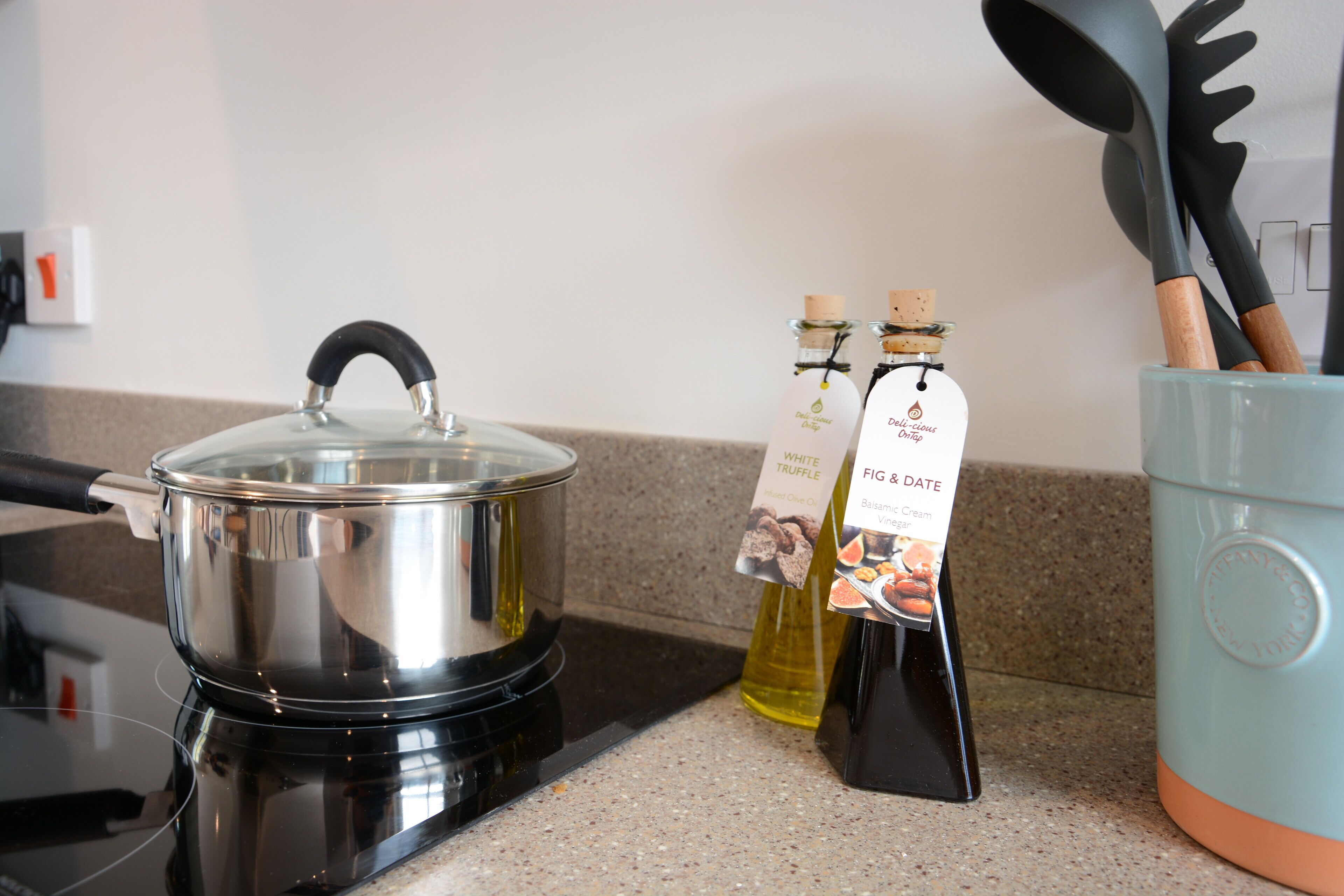 A kitchen with a stainless steel pot on an induction cooktop, two bottles of olive oil, and a blue container holding wooden utensils.