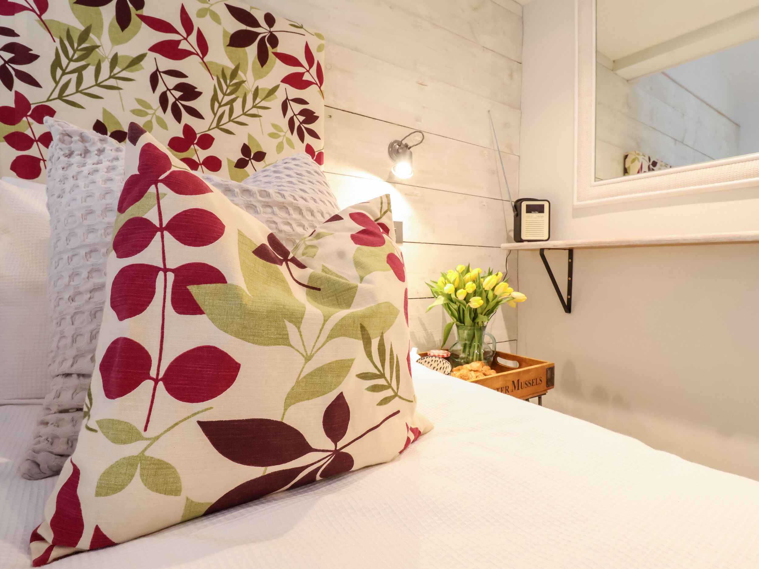 A bedroom with a bed, a pillow with a leaf pattern, a vase with yellow flowers, and a bedside table with a book and a lamp.