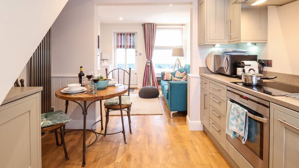 Une cuisine moderne avec un plancher en bois, un coin repas avec une table et des chaises, et une vue sur un salon avec un canapé et des rideaux.