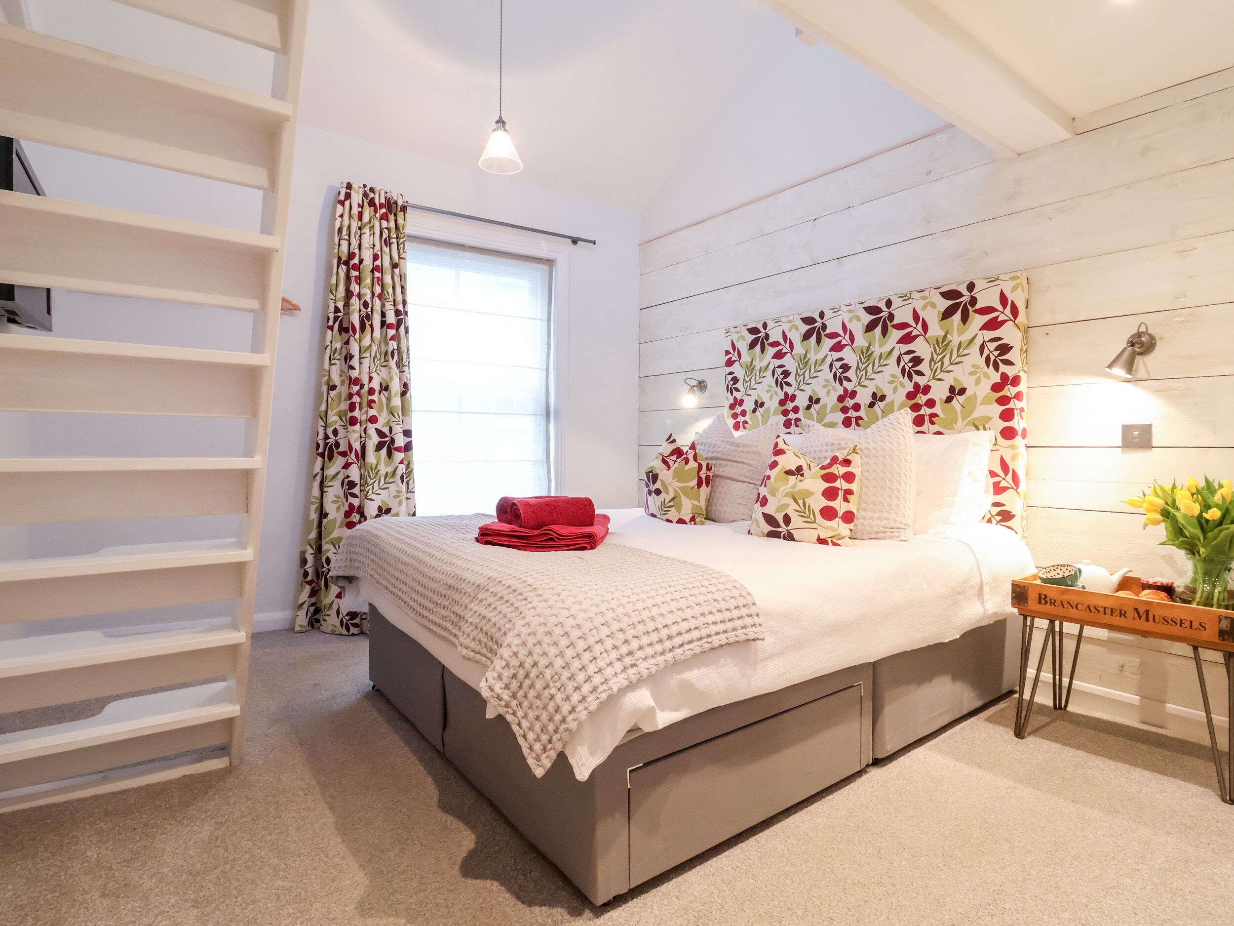 Une chambre à coucher avec un lit, une table de chevet ornée d’un vase, une étagère et une fenêtre avec des rideaux.