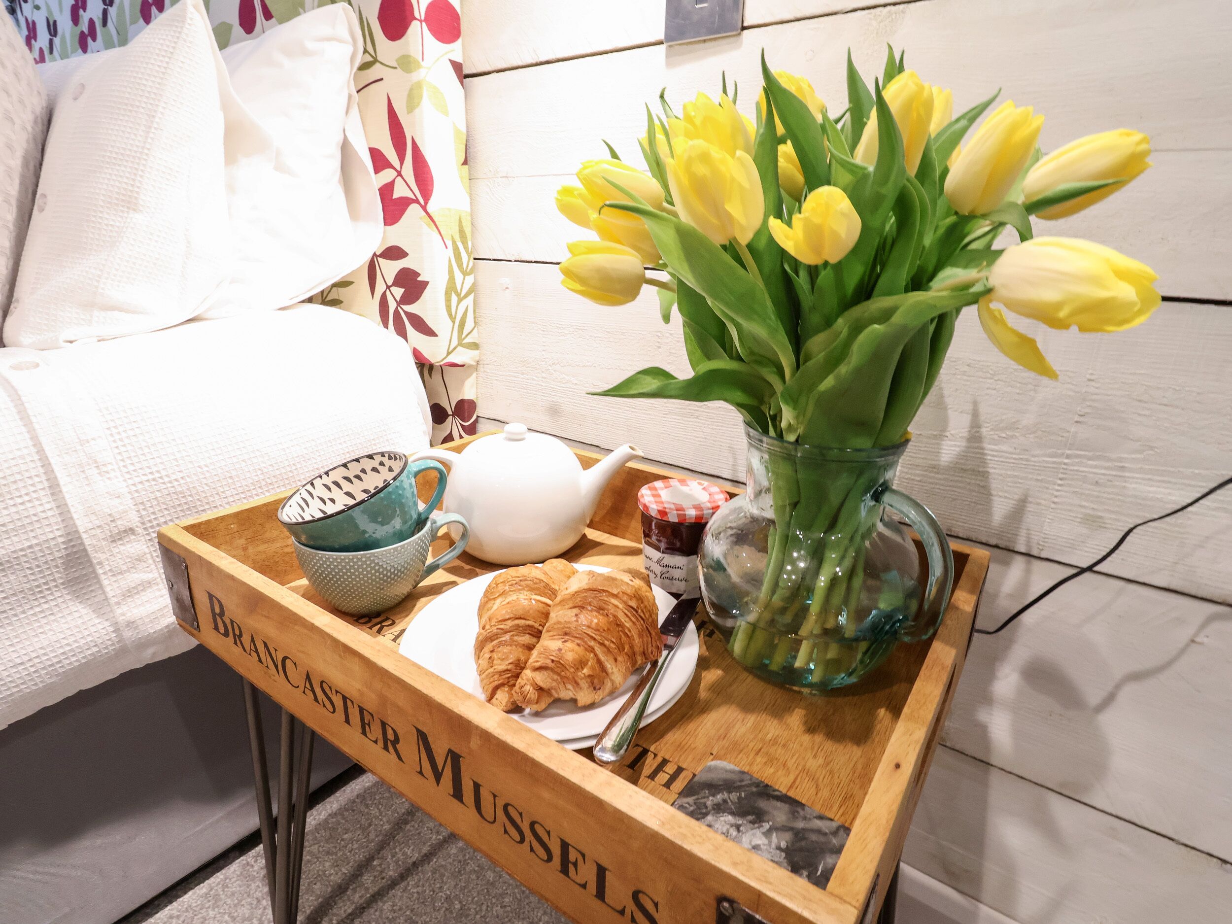 A tray with croissants, a teapot, a mug, a jar of jam, and a vase of yellow tulips on a wooden table.