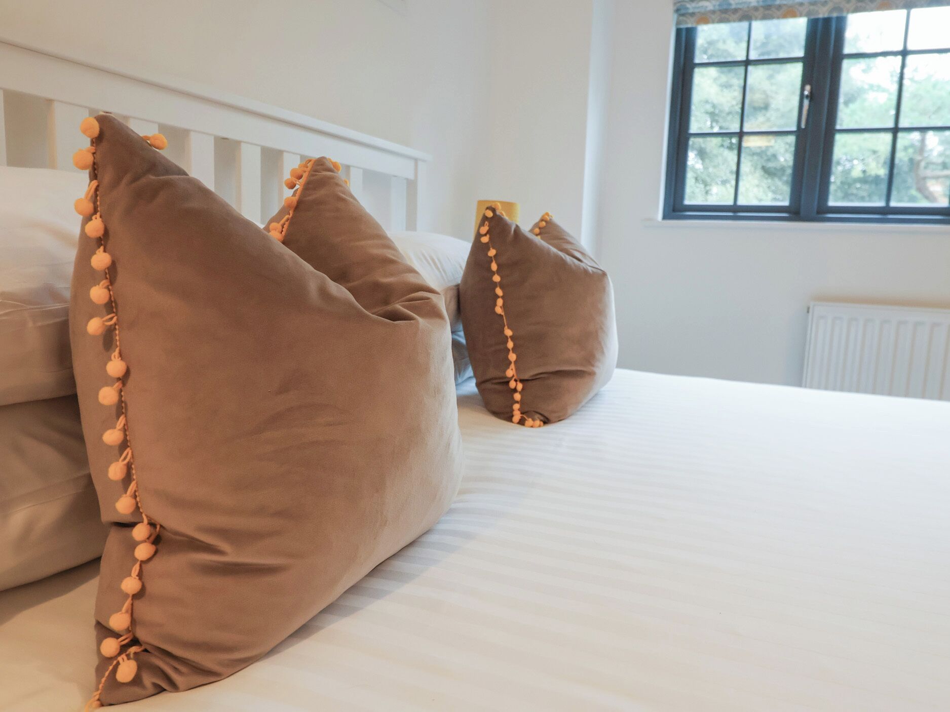 A bed with white bedding and three brown pillows with pom-pom tassels.