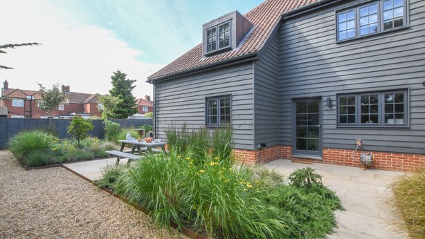 A two-story house with a patio area, a small garden, and a visible chimney. - Corner Cottage, Thorpeness (Leiston)
