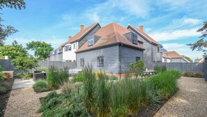 A two-story house with a grey exterior and a red-tiled roof, surrounded by a gravel driveway and a variety of plants. - Corner Cottage, Thorpeness (Leiston)