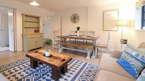 A cozy living room with a wooden coffee table, a dining table set for four, a sofa with patterned cushions, a kitchen area with a wooden cabinet, and a wall clock. - Corner Cottage, Thorpeness (Leiston)
