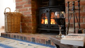 Una chimenea con pared de ladrillo, una cesta de mimbre, una copa de vino tinto y un libro sobre una mesa.