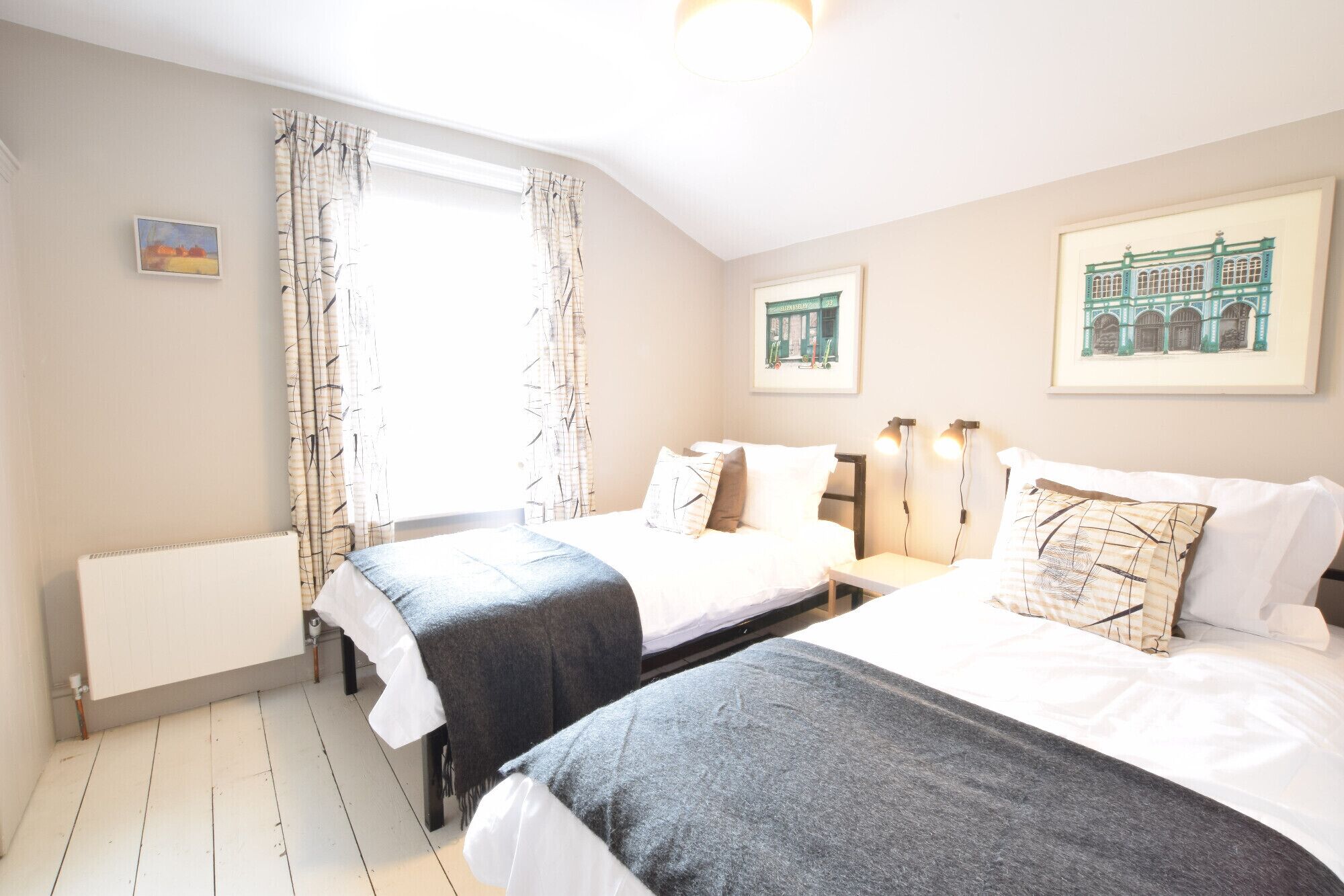 A bedroom with two beds, a radiator, a window with curtains, and framed pictures on the walls.