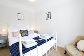 A neatly made bed with a white metal frame, a blue blanket, and a pillow with a peacock design. There are two framed pictures on the wall, a bedside lamp, and a small bedside table with a lamp. - Satis Cottage, Westleton (Saxmundham)