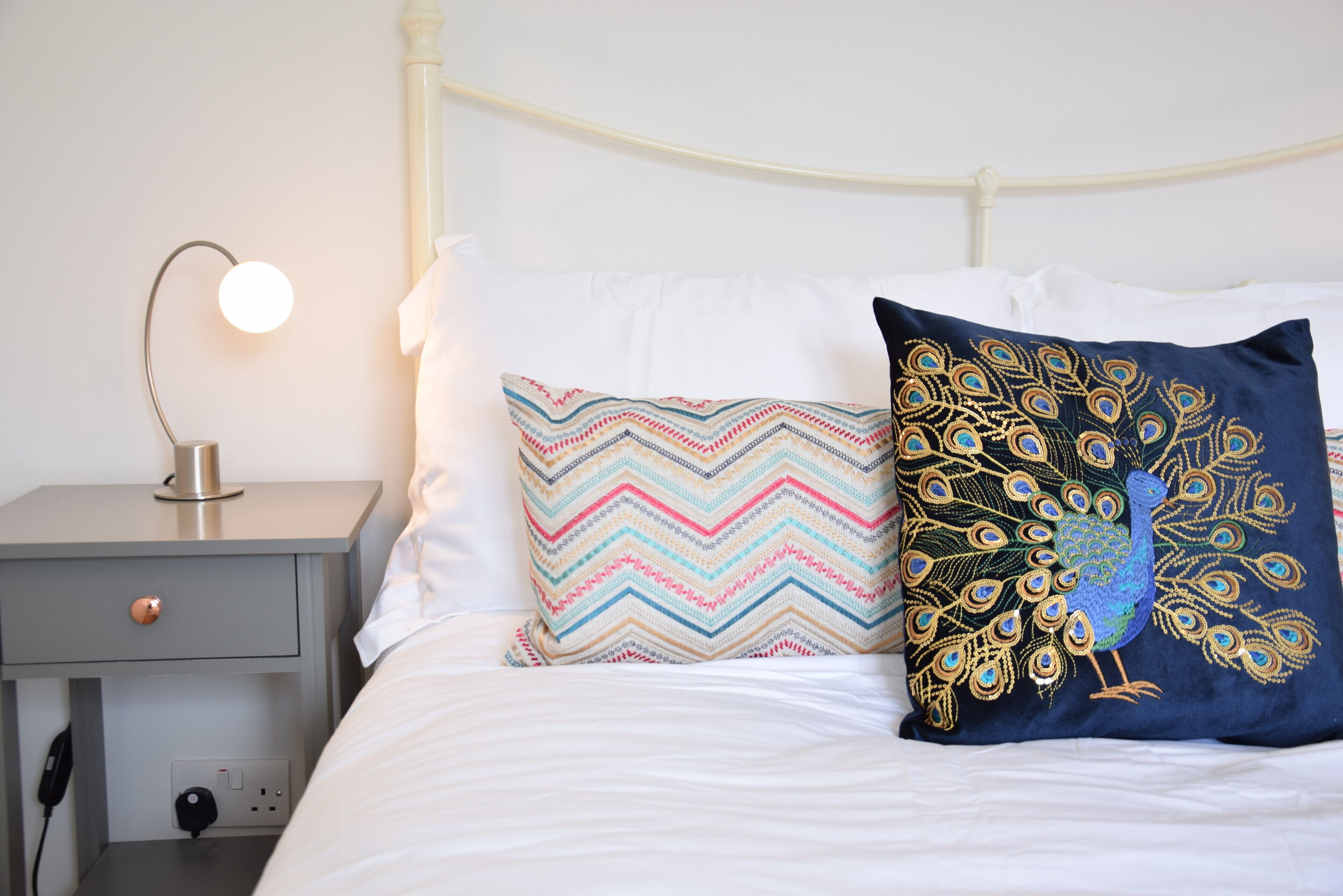 Cama bem arrumada com duas almofadas decorativas, abajur e uma tomada na mesinha de cabeceira.
