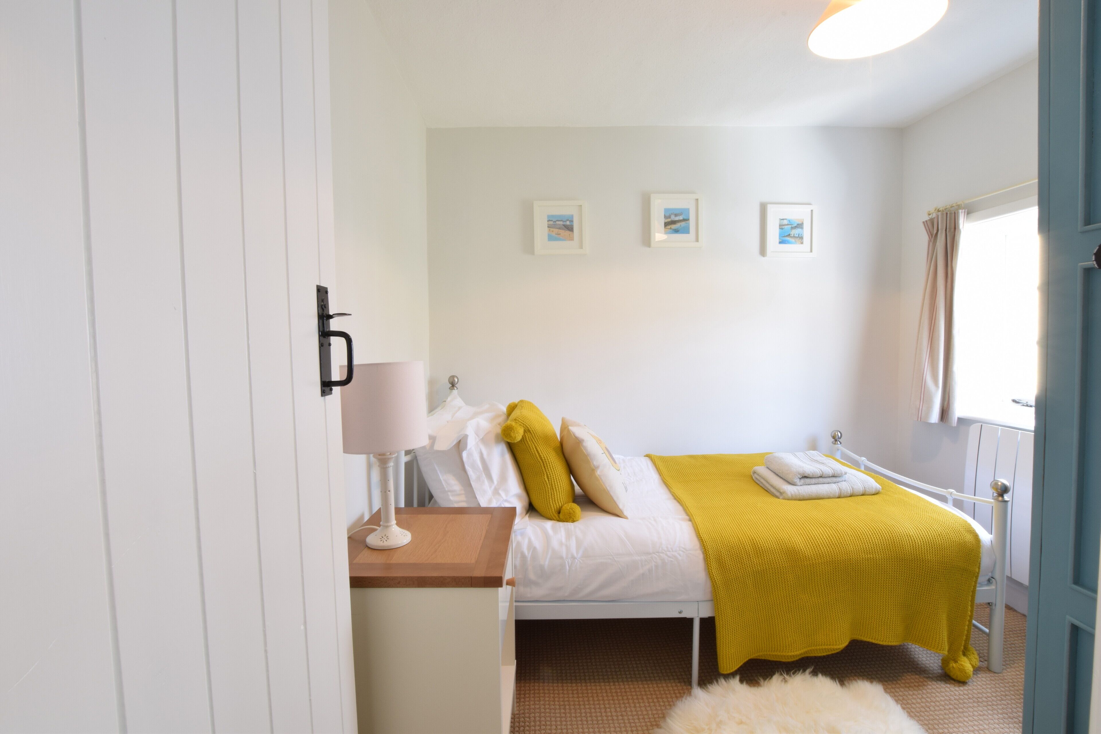 A bedroom with a bed, a bedside table with a lamp, a window with curtains, and three framed pictures on the wall.