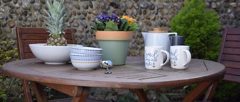 A round wooden table with a pineapple, a potted plant with purple and orange flowers, a white pitcher, and two white mugs with blue text.