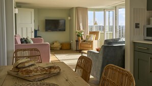 Un vaste salon comprenant une table à manger en bois, des chaises en osier, un canapé et un fauteuil gris. Un téléviseur à écran plat est fixé au mur, et une porte-fenêtre coulissante donne accès à un balcon.