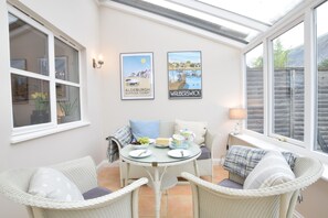 A conservatory with a glass table and wicker chairs, a framed picture of a coastal scene, and a lamp on a window sill. - Bluebelle Cottage, Peasenhall (Saxmundham)