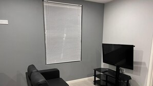 A modern living room with a grey wall, a black leather sofa, a TV on a black stand, and a window with blinds. - Luxury Apartment - Free Wi-fi & Parking NO Deposit (Coventry)