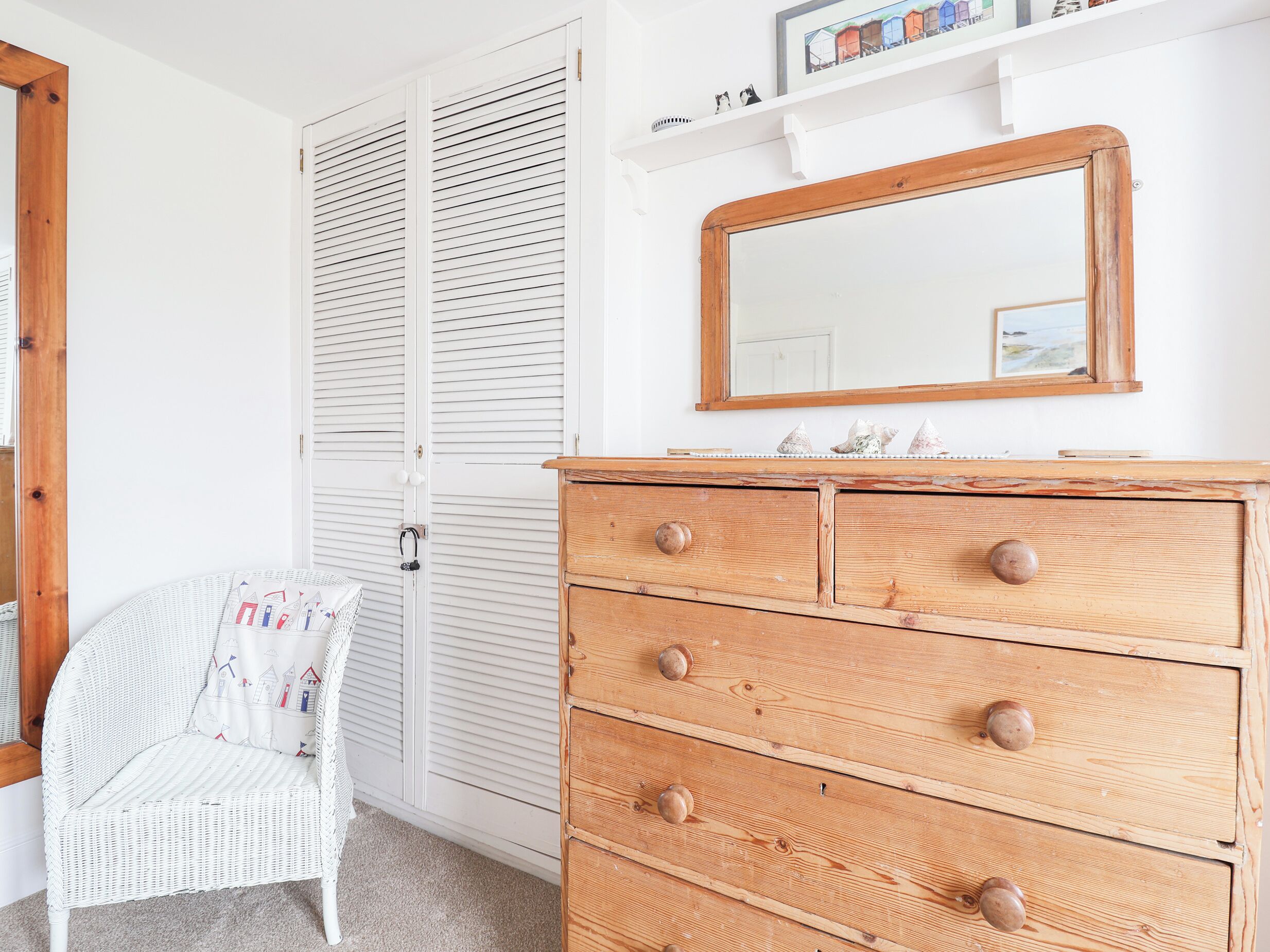 A room with a white wicker chair, a wooden dresser with drawers, a white closet with louvered doors, and a shelf with decorative items.