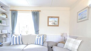 A living room with two sofas, a coffee table, and a side table with a lamp. There is a window with curtains, a framed picture on the wall, and a shelf with decorative items.