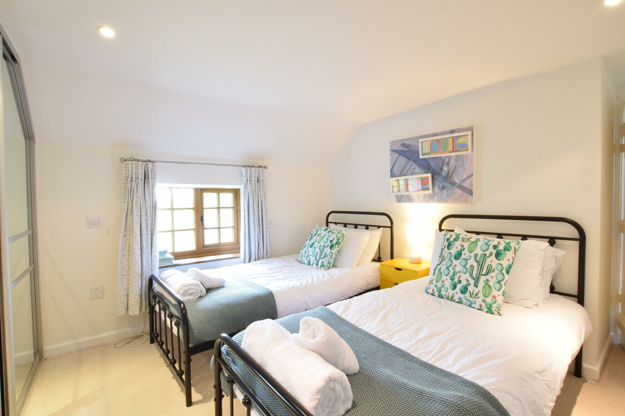 A bedroom with two beds, a window with curtains, a bedside table with a lamp, and a wall art piece.