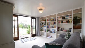 A living room with a grey sofa, a bookshelf filled with books, and a large window with a view of greenery outside. - Skelder, Parham (Woodbridge)