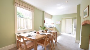 A dining area with a wooden table and chairs, a window with curtains, and a kitchen in the background. - 14 Dunwich Road, Southwold (Reydon)