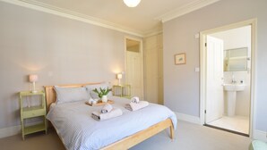 A bedroom with a bed, bedside tables, a nightstand, a bedside lamp, a potted plant, and a bathroom visible through an open door. - 14 Dunwich Road, Southwold (Reydon)