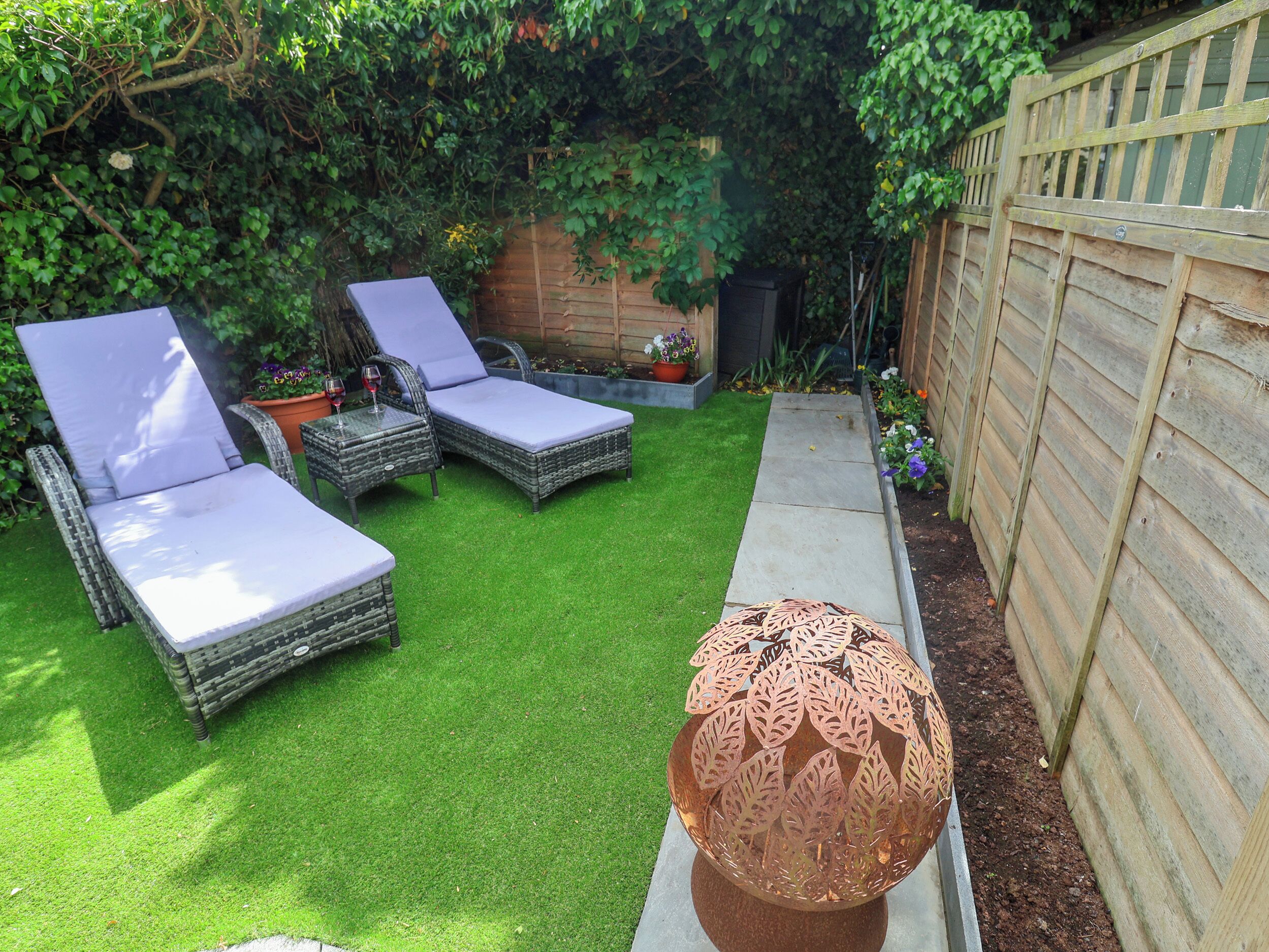 A backyard with artificial grass, two wicker lounge chairs, a decorative metal sphere, and a wooden fence.