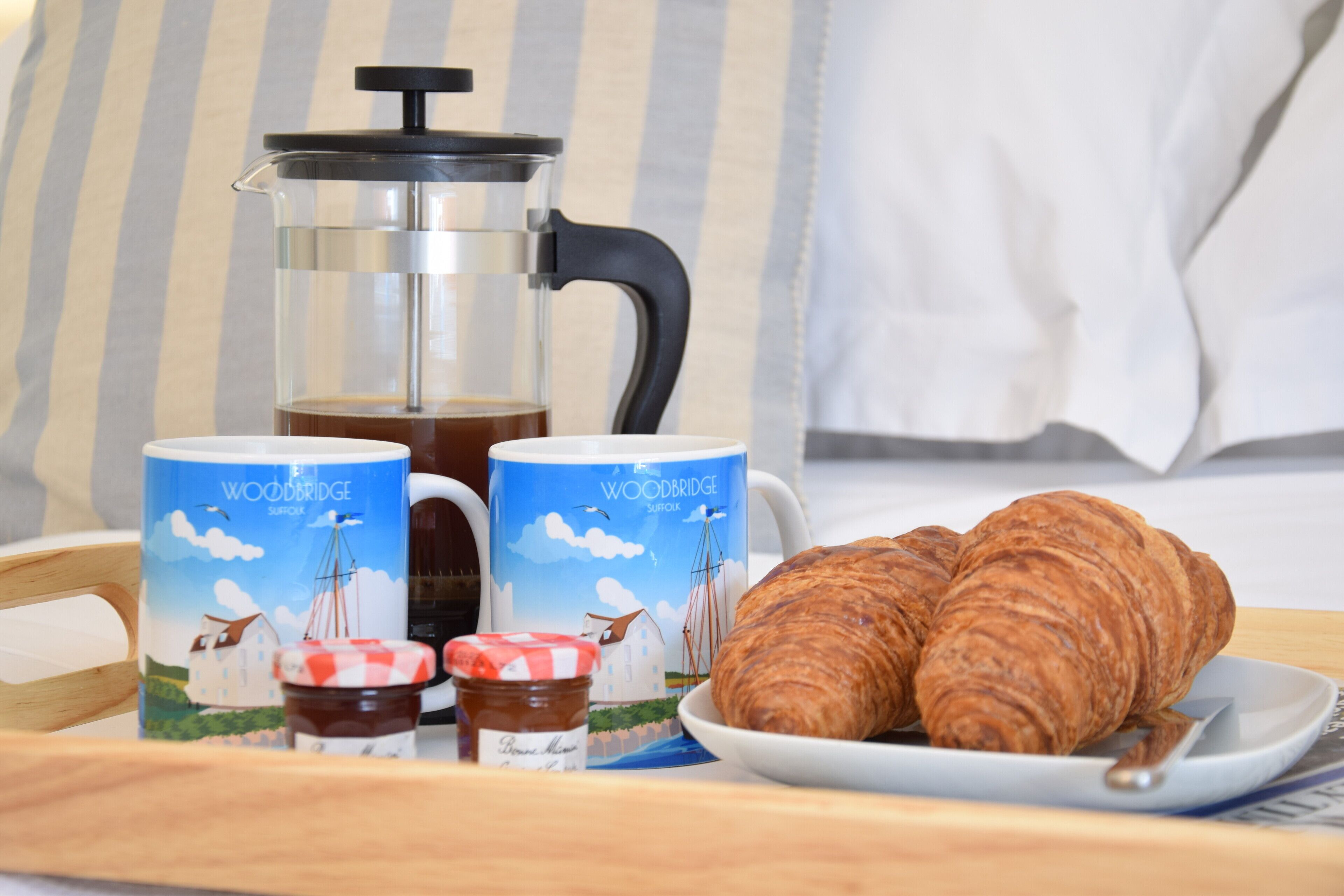 A breakfast setup with a French press, two mugs, a jar of jam, and two croissants on a tray.