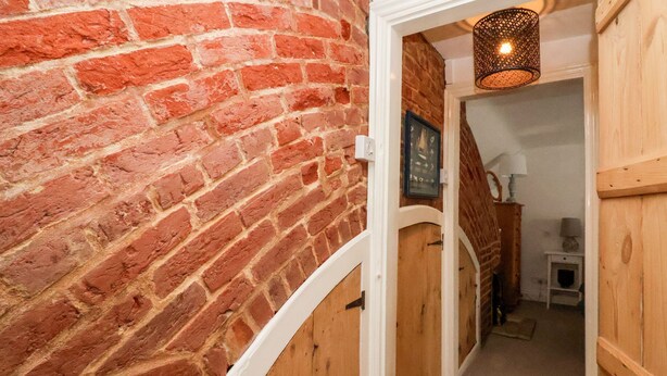 A brick wall with a wooden door leading to a room with a lamp and a small table. - Cobblers Cottage, Southwold (Reydon)