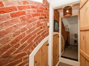 A brick wall with a wooden door leading to a room with a lamp and a small table. - Cobblers Cottage, Southwold (Reydon)