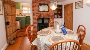 A dining area with a table set for four, surrounded by wooden chairs. A brick fireplace is visible in the background, and a kitchen is partially seen to the left. - Cobblers Cottage, Southwold (Reydon)
