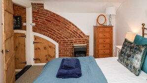 A bedroom with a brick archway, a bed with a blue blanket, a wooden dresser, and a nightstand with a lamp. - Cobblers Cottage, Southwold (Reydon)