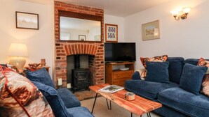 A living room with a brick fireplace, a television, and blue sofas. - Cobblers Cottage, Southwold (Reydon)