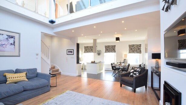 Ein großzügiger Wohnbereich mit grauem Sofa, Couchtisch und Kamin. Es gibt einen Essbereich mit Tisch und Stühlen, und im Hintergrund eine Küche.