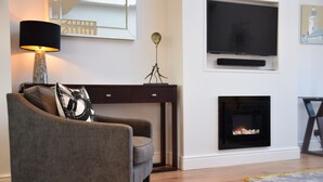 Ein modernes Wohnzimmer mit grauem Sessel, einer schwarzen Lampe, einem Kamin, einem Flachbildfernseher und einem Konsolentisch.